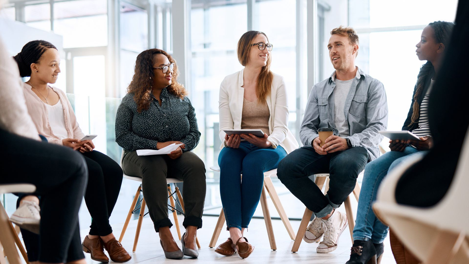 Group of six diverse adults sitting in a circle in a modern, well-lit room having a discussion, some holding notepads and tablets, participating in a collaborative session or group therapy.