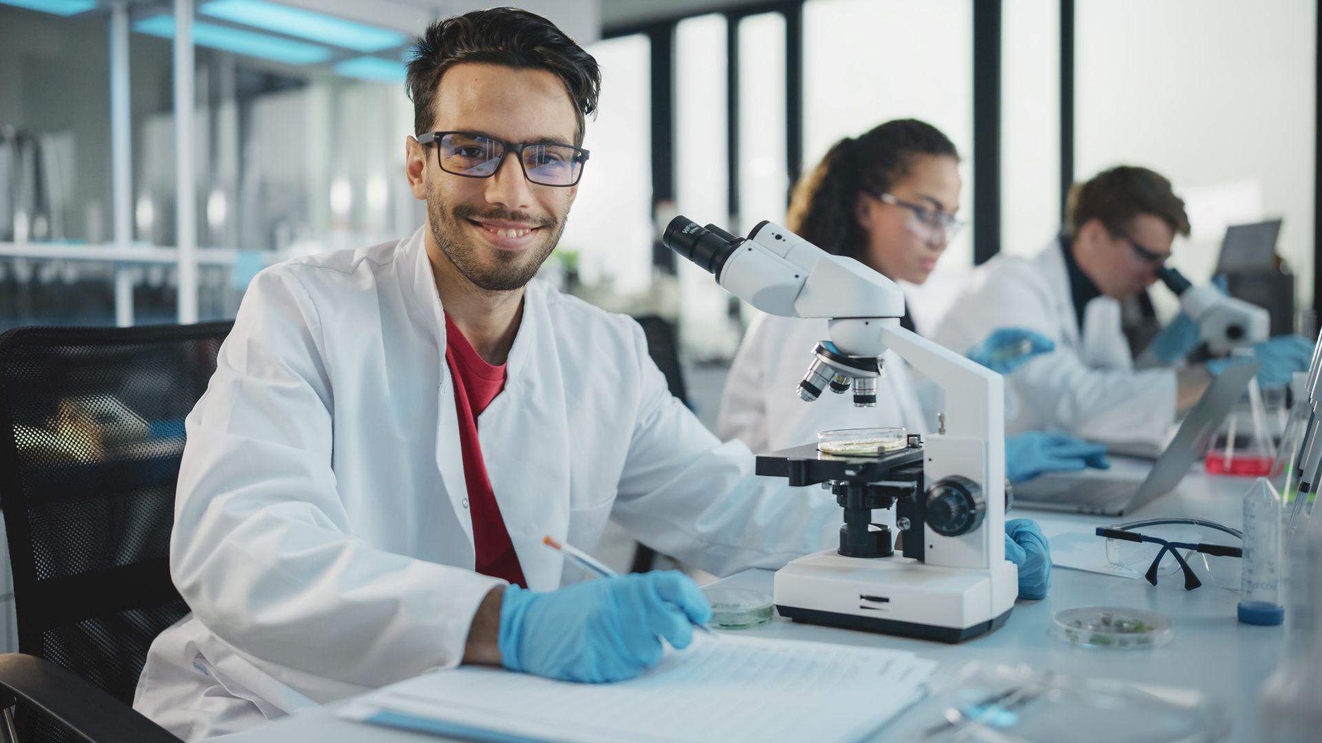 Smiling researcher in a white lab coat and gloves working at a microscope, with lab notes open in front of him; two colleagues in the background also conducting experiments.