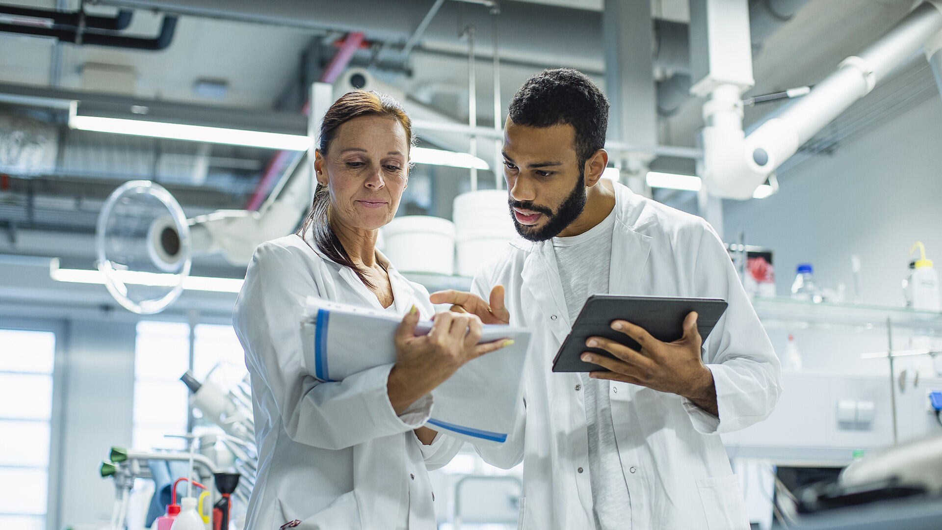 Two scientists in lab coats collaborating in a modern laboratory, with one holding a tablet and the other reviewing documents as they discuss research data.