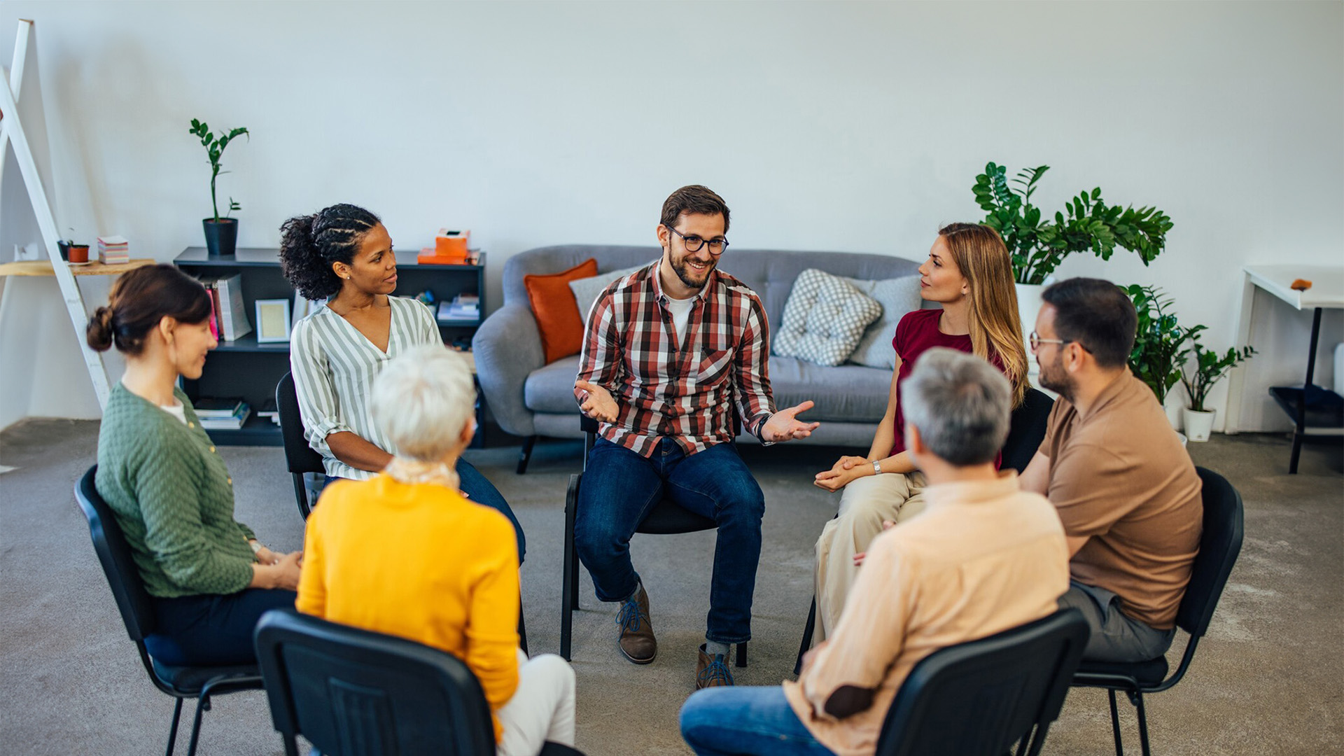 A diverse group of people sitting in a circle during a group therapy or support session, with one man speaking while others listen attentively in a comfortable, casual setting.